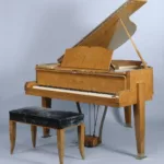 Piano à queue Gaveau du salon de musique de première classe du paquebot Liberté, réalisé vers 1950 en bouleau de Norvège, avec couvercle relevé, pupitre intégré, pédalier apparent et pieds terminés par des sabots en bronze doré, accompagné de son tabouret en velours sombre.