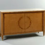 Meuble d’appui du salon de musique de première classe du paquebot Liberté, réalisé vers 1950 en bouleau de Norvège, formant un buffet bas à deux portes, décoré d’une couronne végétale marquetée et de fausses colonnes d’angle terminées par des sabots métalliques, avec plateau en marbre clair.