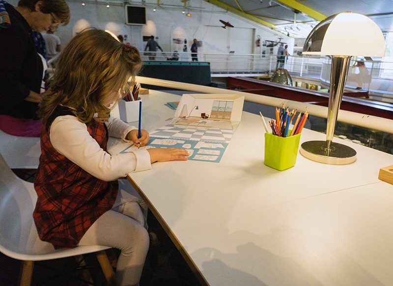 Petite fille coloriant sa planche de jeu "Créez votre intérieur" sur la mezzanine de l'Écomusée de Saint-Nazaire.