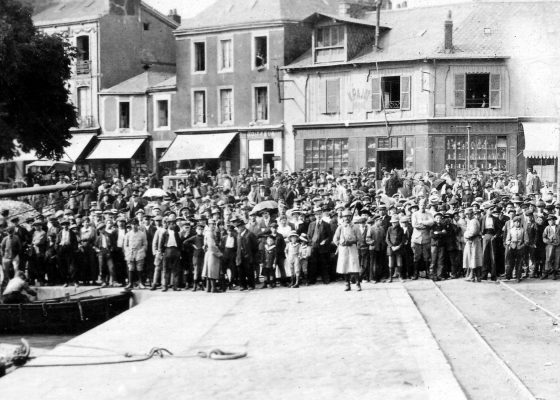 Foule de Nazairiens massée au bout d'un quai retenue par des gendarmes et assistant avec curiosité au débarquement des premiers soldats américains dans le port de Saint-Nazaire pendant la Première Guerre mondiale.