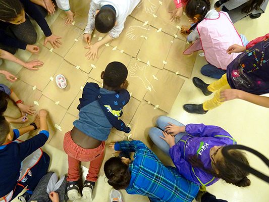 Jeunes élèves de primaire vus du dessus en train de dessiner le contour de leur main sur des cartons au cours d'une action pédagogique.