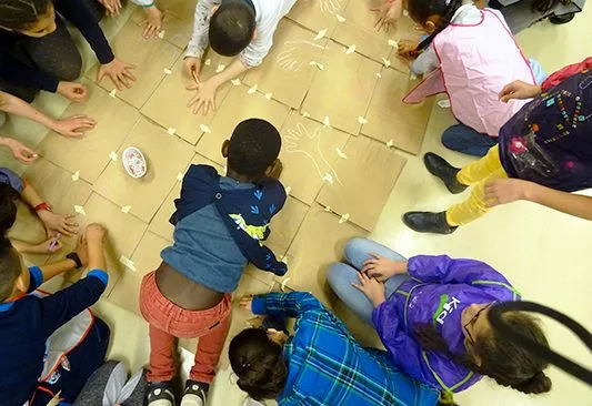 Photographe : Mireille Petit Jeunes élèves de primaire vus du dessus en train de dessiner le contour de leur main sur des cartons au cours d'une action pédagogique.