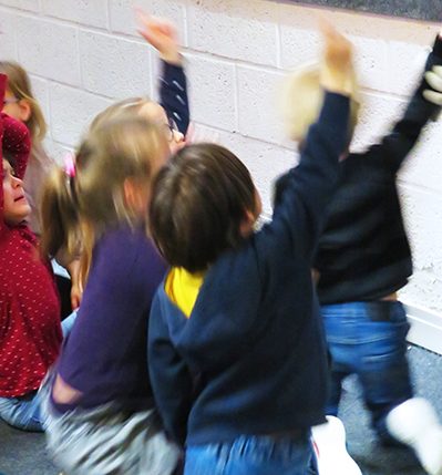 Élèves de maternelle assis de dos en train de lever le doigt pendant une visite scolaire dans l'Écomusée, musée d'histoire de Saint-Nazaire.