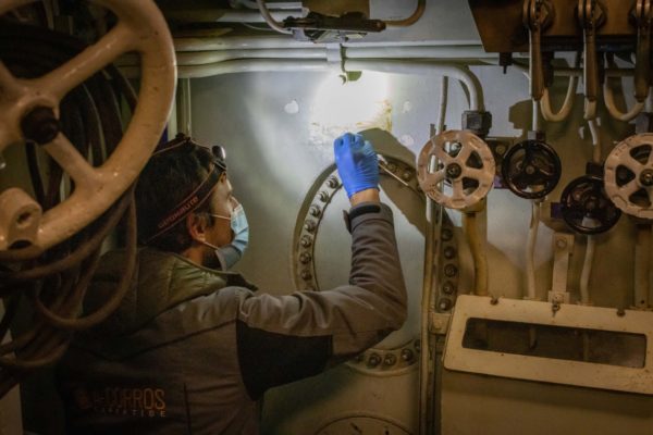 Un homme, équipé d'une lampe frontale et d'une combinaison, applique un produit au coton-tige sur une surface intérieure abîmée du sous-marin Espadon