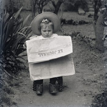 Fillette avec un chapeau lisant le journal sur un petit siège dans une allée de jardin.
