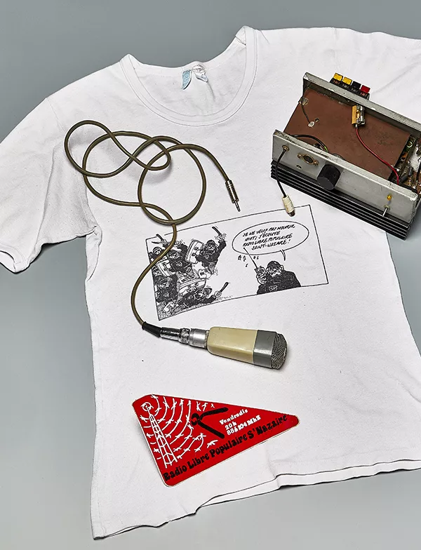 (Tee-shirt) Exécutant : Régate. (Micro) Exécutant : LEM. (Autres) Exécutant inconnu. Collection Saint-Nazaire Agglomération Tourisme – Écomusée. Cliché : Jean-Claude Lemée Tee-shirt blanc sur lequel sont posés un émetteur radio, un micro et un autocollant de la Radio Libre Populaire de Saint-Nazaire.