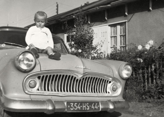 Jeune enfant assis sur le capot d’une voiture de marque Simca devant un baraquement provisoire dans les années 1950.