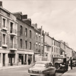 Photographie en noir et blanc de voitures des années 1950 garées dans une rue de Saint-Nazaire dont deux cafés à identifier.