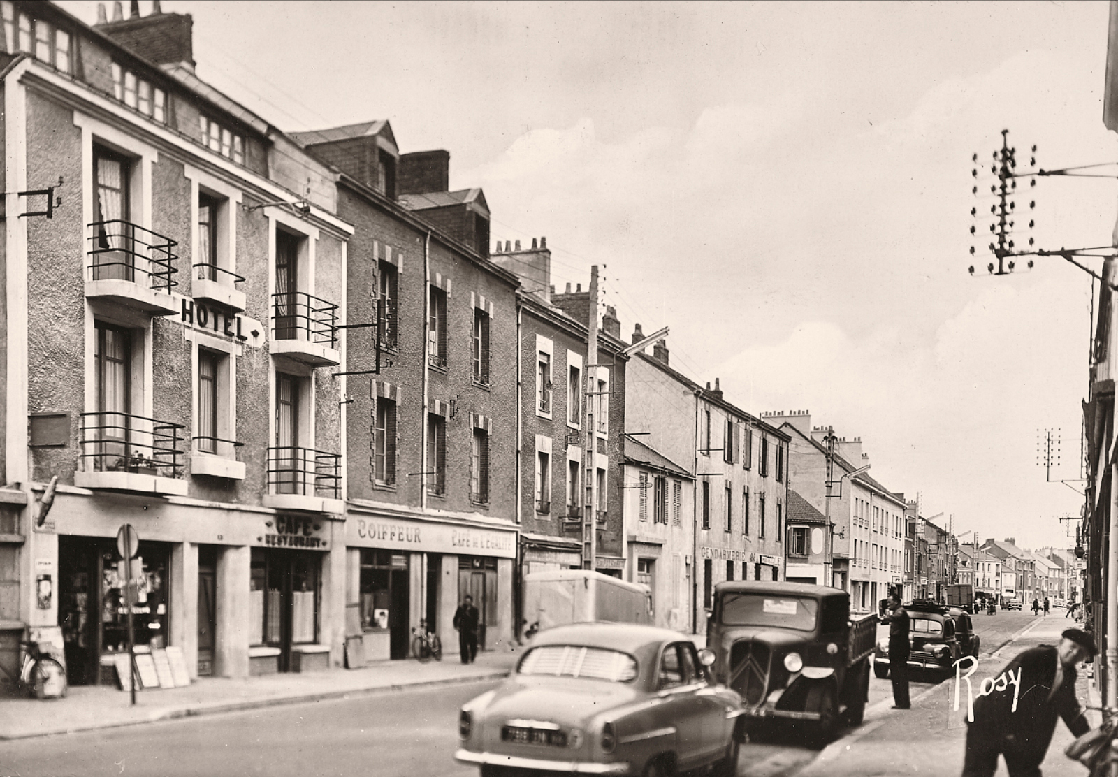 Photographie en noir et blanc de voitures des années 1950 garées dans une rue de Saint-Nazaire dont deux cafés à identifier.