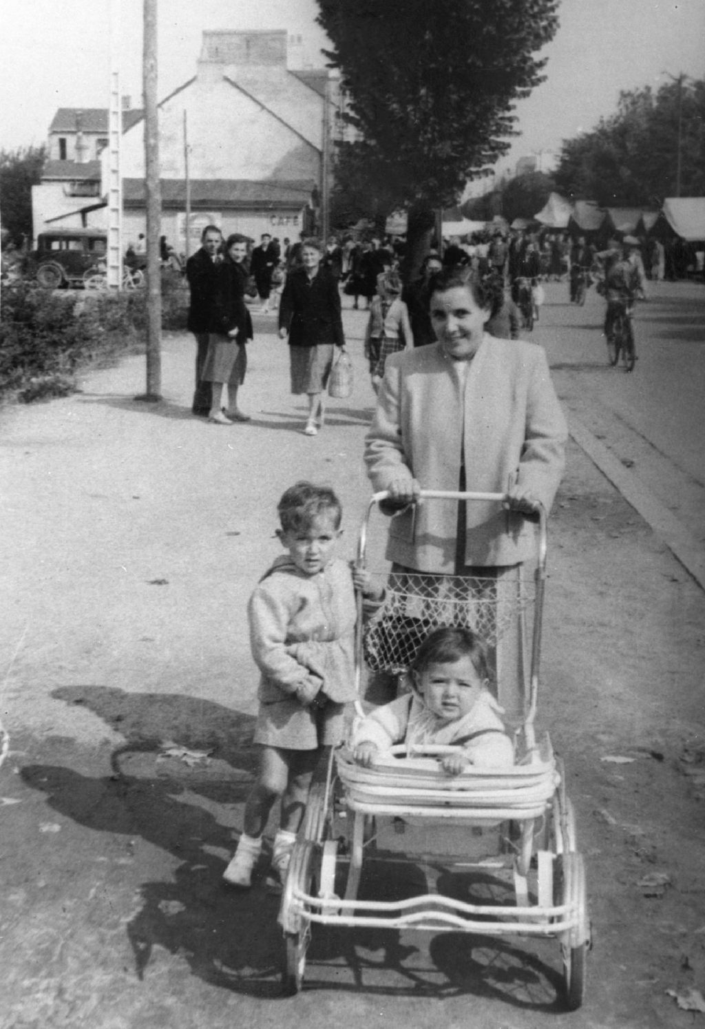 Photographie en noir et blanc représentant une femme avec deux enfants dont un en poussette, à l'arrière-plan des gens marchent dans la rue.
