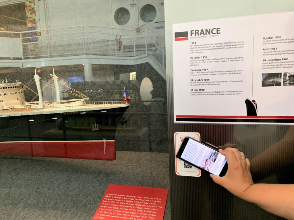 Main tenant un smartphone devant un QR code à côté d'une maquette du paquebot France (1962) à l'Écomusée.