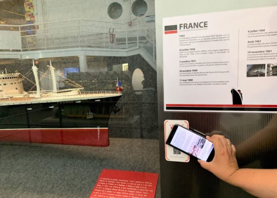 Main tenant un smartphone devant un QR code à côté d'une maquette du paquebot France (1962) à l'Écomusée.