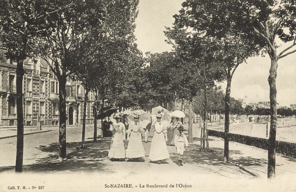 Trois femmes en robe de dentelle blanche et ombrelle se baladent face à nous sur le front de mer vers 1910.