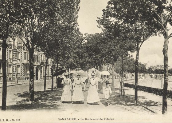 Trois femmes en robe de dentelle blanche et ombrelle se baladent face à nous sur le front de mer vers 1910.