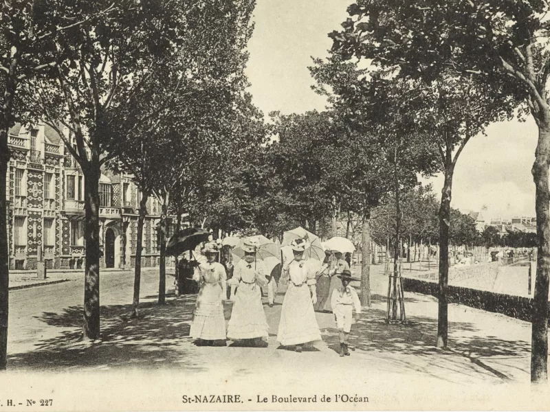 Photographe inconnu. Éditeur : T.H. Collection Saint-Nazaire Agglomération Tourisme Trois femmes en robe de dentelle blanche et ombrelle se baladent face à nous sur le front de mer vers 1910.