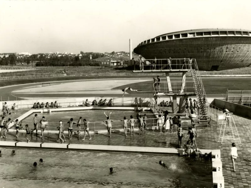 Photographe : Paul Lemasson. Architectes : Louis Longuet, René Rivière et Roger Vissuzaine. Collection Saint-Nazaire Agglomération Tourisme-Écomusée Photographie noir et blanc représentant une vue aérienne de deux bassins extérieurs de la piscine Léo Lagrange avec de nombreux baigneurs devant la Soucoupe.