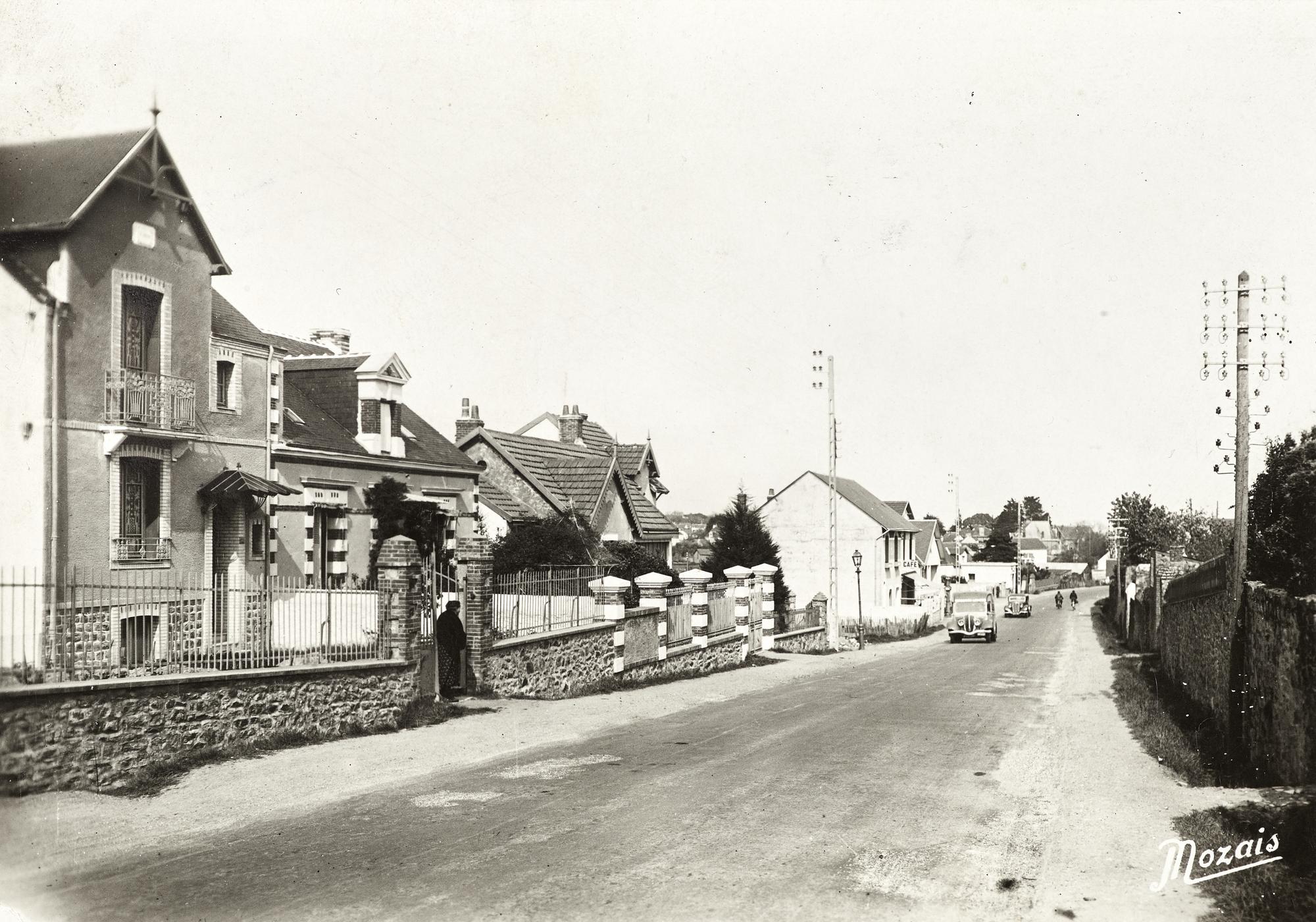 Carte postale noir et blanc représentant une rue en pente de Saint-Nazaire où circulent deux voitures dans les années 1930.