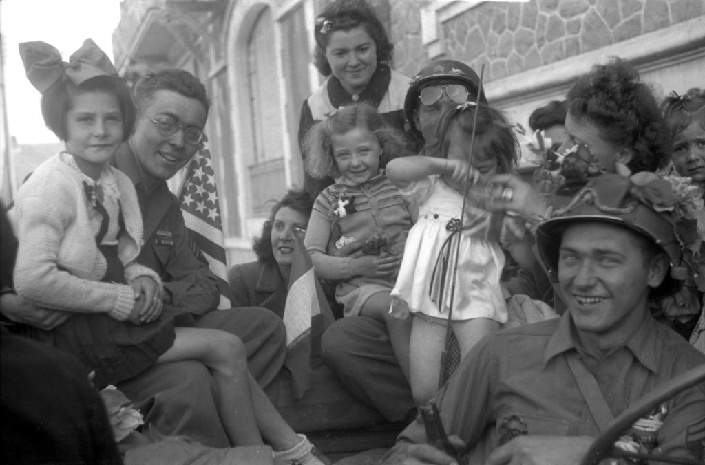 Photographie en noir et blanc représentant des soldats américains posant sur une jeep avec des enfants et des jeunes femmes à la Libération.