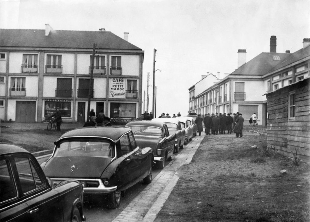 Photographie noir et blanc d'une file de voitures garées le long d'une rue dans le quartier du Petit Maroc, au fond un groupe d'hommes et un café.