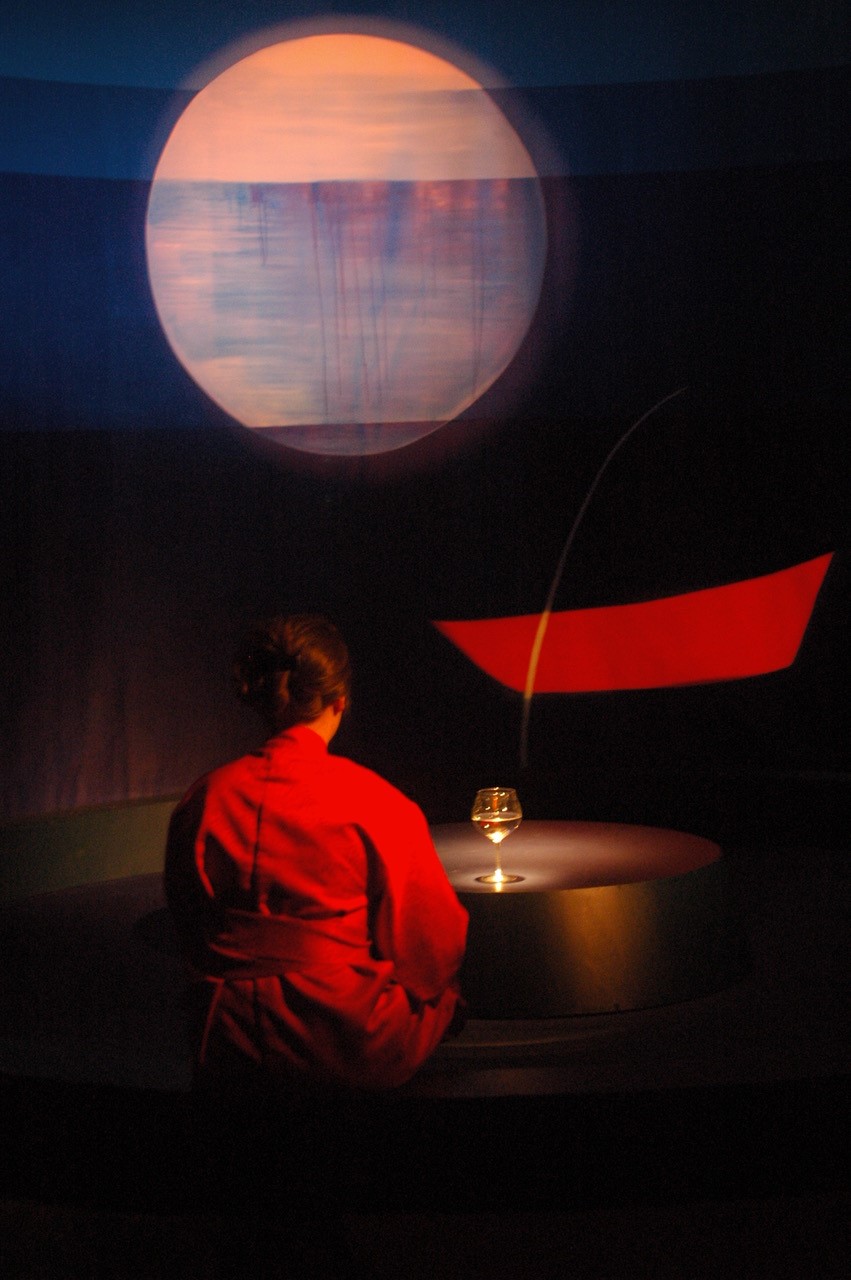 Photographie d'un spectacle musical représentant une jeune femme vêtue de rouge et assise en tailleur dos au spectateur dans une ambiance sombre.