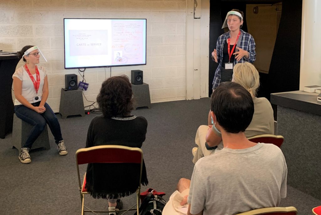 Jeune femme, accompagnée d'une professionnelle de musée, debout avec une visière s'adressant à trois personnes assises devant elle lors de "Ma solidarité" aux Journées du Patrimoine.