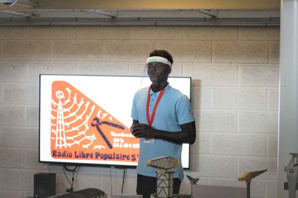 Jeune homme debout avec une visière devant un écran projetant un autocollant de la Radio Libre Populaire lors de "Ma solidarité" aux Journée du patrimoine.