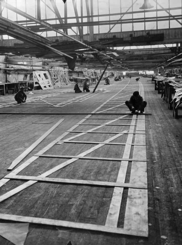 Photographie noir et blanc de la salle à tracer des chantiers navals nazairiens avec des ouvriers accroupis en train de tracer des formes au sol.