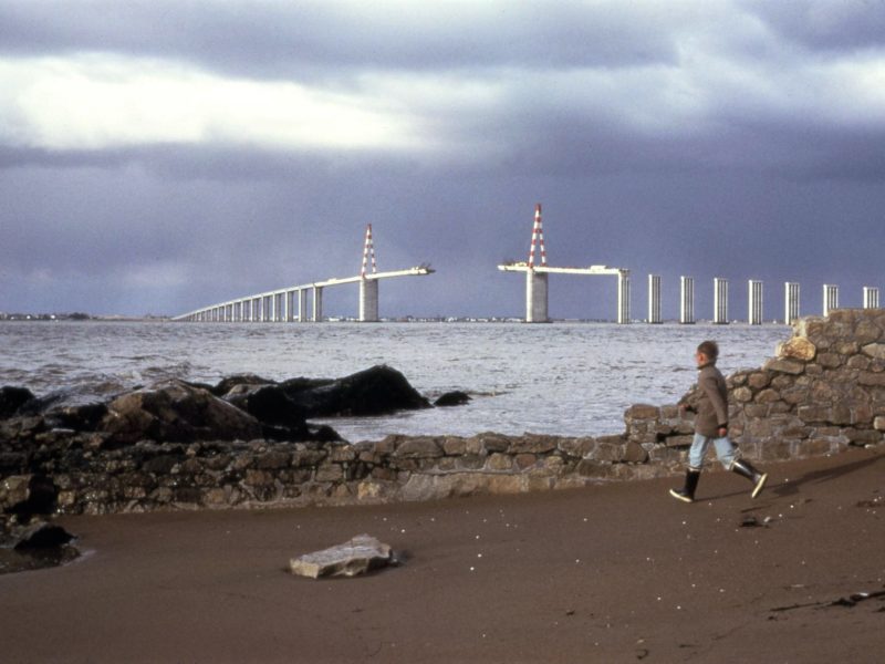 Photographe : Émile Arteaga. Collection Saint-Nazaire Agglomération Tourisme - Écomusée Photographie d'un enfant courant sur une plage devant un muret, derrière l'estuaire de la Loire et le pont de Saint-Nazaire en cours de construction.