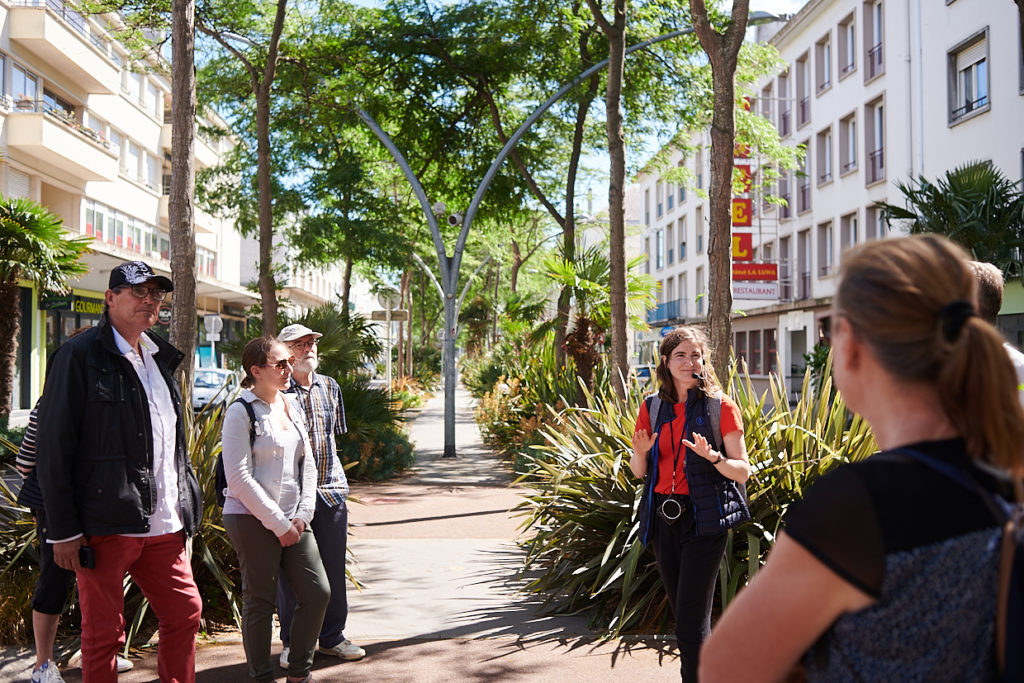 Guide et groupe de visiteurs sur un trottoir sous les arbres et devant les façades d'immeubles de la Reconstruction dans l'avenue de la République à Saint-Nazaire.