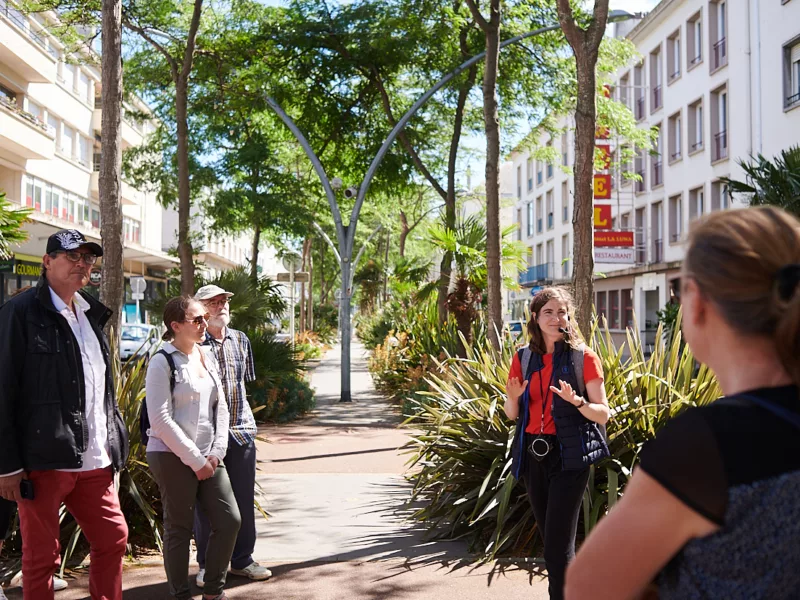 Photographe : Rudy Burbant. Guide et groupe de visiteurs sur un trottoir sous les arbres et devant les façades d'immeubles de la Reconstruction dans l'avenue de la République à Saint-Nazaire.