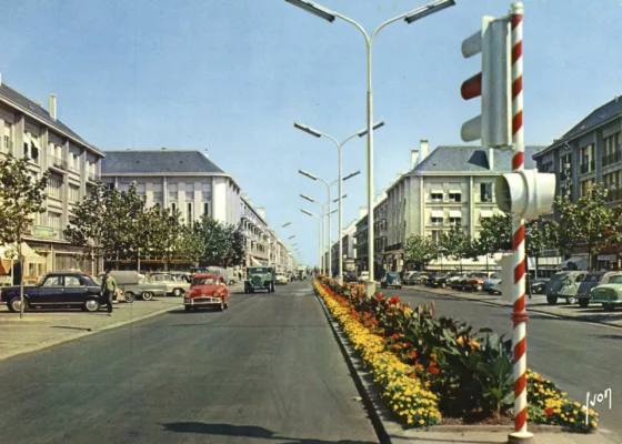 Voitures et camion roulant sur l'avenue de la République pavoisée dans les années 1950 au moment de la Reconstruction.