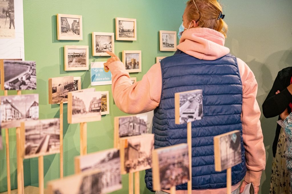 Femme montrant du doigt une carte postale encadrée au mur dans l'exposition "Regard miroir ou le paysage réinventé" à l'Écomusée de Saint-Nazaire