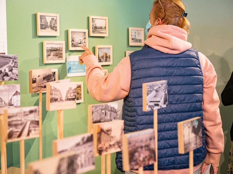 Femme montrant du doigt une carte postale encadrée au mur dans l'exposition "Regard miroir ou le paysage réinventé" à l'Écomusée de Saint-Nazaire