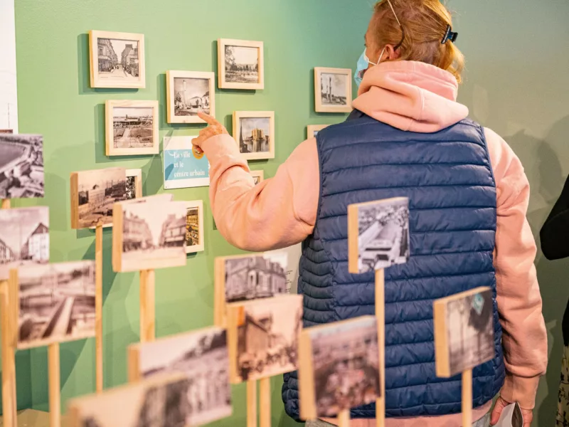 Photographe : Maelwenn Leduc - Saint-Nazaire Agglomération Tourisme Femme montrant du doigt une carte postale encadrée au mur dans l'exposition "Regard miroir ou le paysage réinventé" à l'Écomusée de Saint-Nazaire
