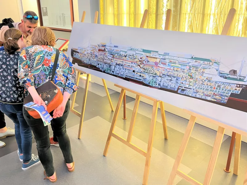 Photographe : Amélie Vincent-Le Berre. Saint-Nazaire Agglomération Tourisme. Famille de deux adultes et une enfant jouant au jeu France a besoin de vous devant un panneau représentant une coupe du paquebot France à Escal'Atlantic.