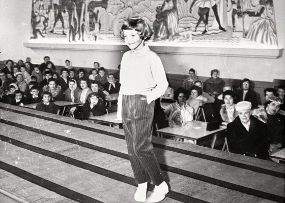 Photographie en noir et blanc représentant une petite fille défilant devant une foule de spectateurs dans la salle de la Mutualité.