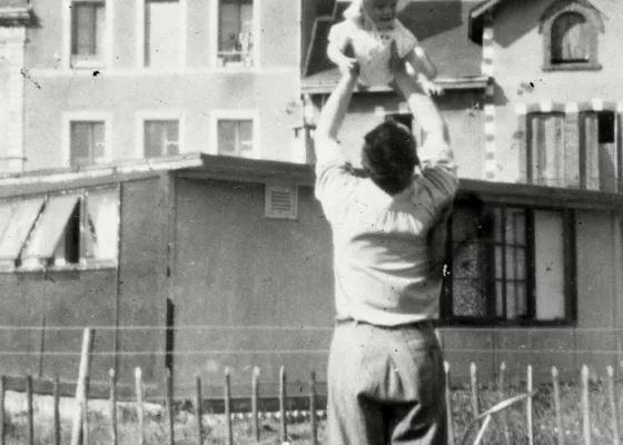Photographie en noir et blanc représentant un homme portant dans les bras son enfant devant un bungalow pendant la reconstruction à Saint-Nazaire