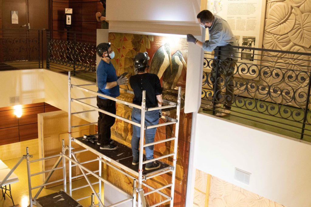 Deux professionnels de musée sont sur un échafaudage pour observer des panneaux de laque accrochés dans le hall d'Escal'Atlantic.