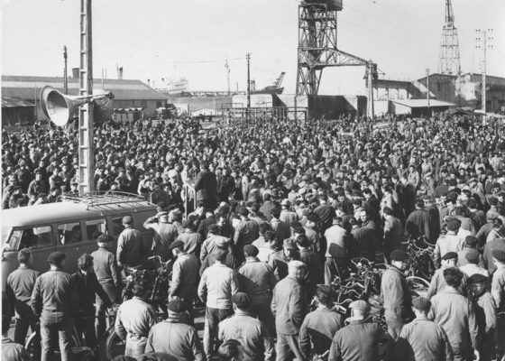 Un homme fait un discours au milieu d'une foule d'ouvriers aux chantiers navals lors des grèves de 1957.