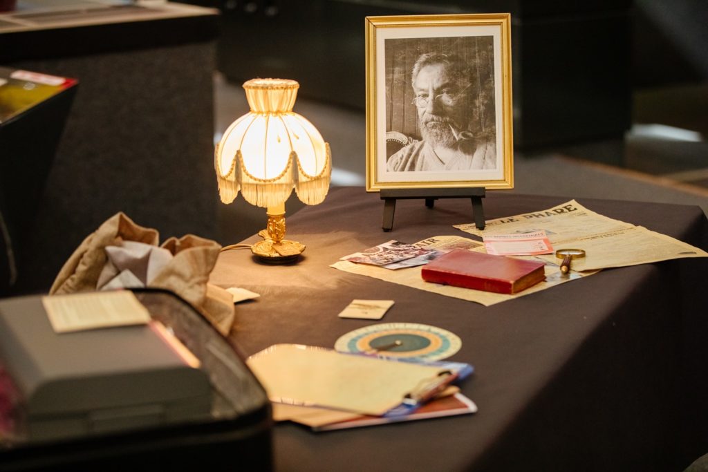Table recouverte d'objets mystérieux et du portrait photographique d'un homme âgé éclairés par une lampe de chevet ancienne.