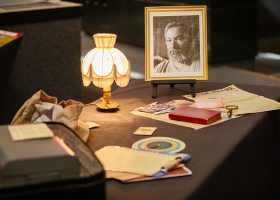 Table recouverte d'objets mystérieux et du portrait photographique d'un homme âgé éclairés par une lampe de chevet ancienne.