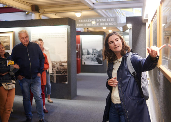 Photographe : Rudy Burbant. Guide montrant une œuvre à un groupe de visiteurs pendant une visite de l'Écomusée.