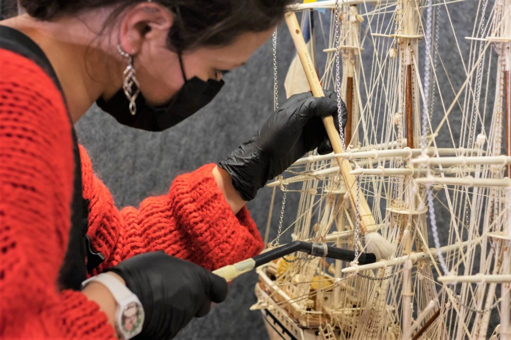 Professionnelle de musée dépoussiérant une maquette du quatre-mâts Tarapaca à l'aide d'un pinceau dans l'Écomusée.