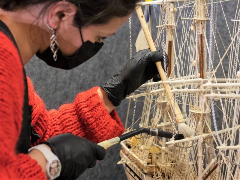 Professionnelle de musée dépoussiérant une maquette du quatre-mâts Tarapaca à l'aide d'un pinceau dans l'Écomusée.