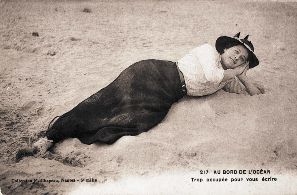 Carte postale souvenir représentant une jeune femme allongée sur la plage. La légende de la carte indique : "au bord de l'océan, trop occupée pour vous écrire".