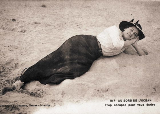 Carte postale souvenir représentant une jeune femme allongée sur la plage. La légende de la carte indique : "au bord de l'océan, trop occupée pour vous écrire".