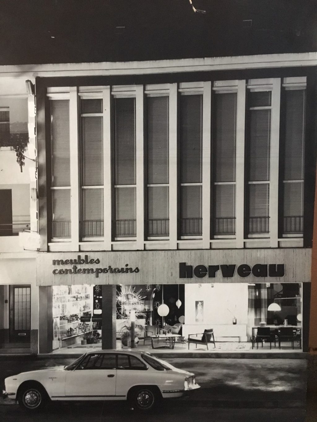 Façade d'un immeuble de la Reconstruction la nuit avec la vitrine du commerce de meubles Herveau allumée au rez-de-chaussée.