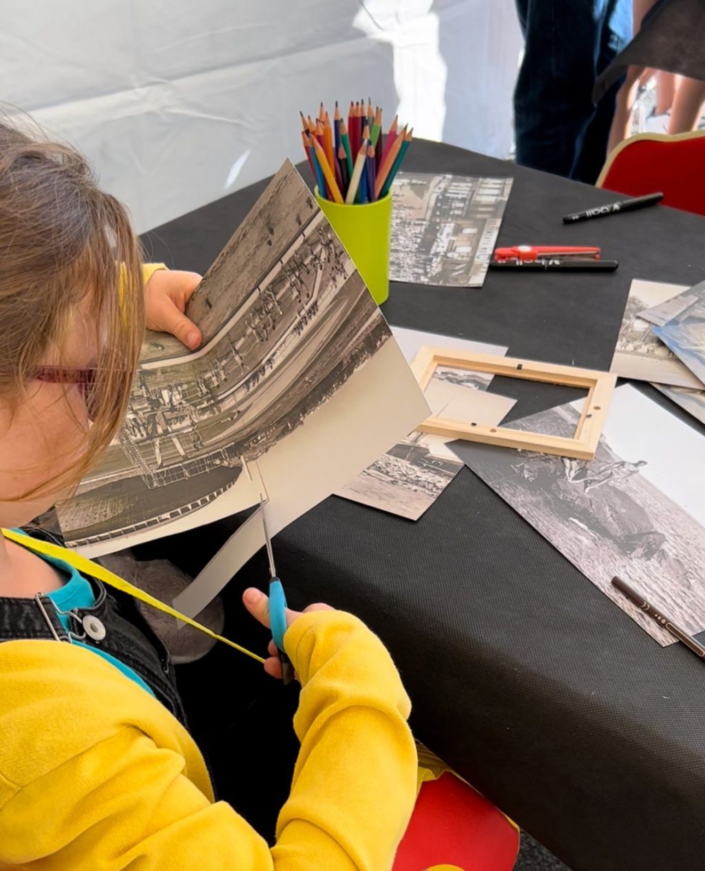 Une enfant de dos découpe une photographie pour créer sa carte postale lors d'un atelier à l'Écomusée.