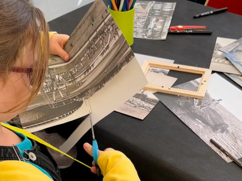 Une enfant de dos découpe une photographie pour créer sa carte postale lors d'un atelier à l'Écomusée.