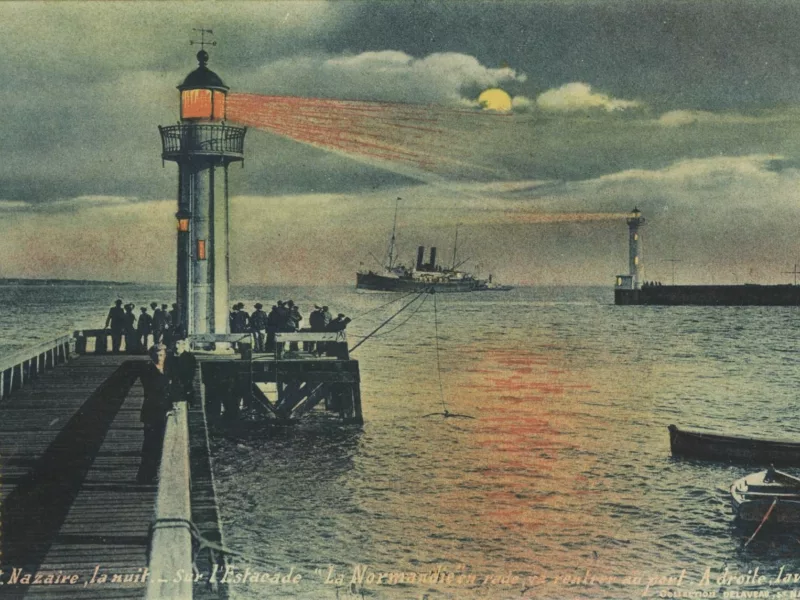 Photographe : inconnu. Éditeur : Delaveau. Collection Saint-Nazaire Agglomération Tourisme - Écomusée. Carte postale illustrée représentant l'une des estacades de l'entrée est et le vieux môle de nuit, au loin un paquebot s'avance alors que le feu de l'estacade projette une lumière orangée sur la mer.